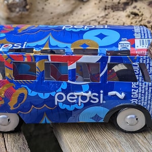 May include: A blue and gold toy bus made from recycled aluminium cans, featuring the Pepsi logo. The bus has a floral pattern and the words "pepsi" and "có gaz peg ula" printed on the side.