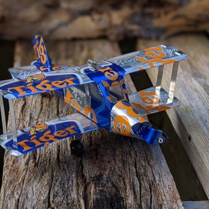 Peut inclure: Un modèle de biplan artisanal fabriqué à partir de canettes de bière Tiger recyclées. L'avion jouet présente des couleurs bleues, oranges et argentées, avec le logo "Tiger" visible sur les ailes et le fuselage. Le modèle est posé sur un fond en bois rustique.