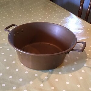 Puede incluir: Una olla de cocina de metal marrón con dos asas laterales. La olla es redonda y tiene una abertura ancha. Está sobre un mantel verde claro con lunares blancos.