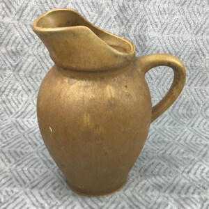 May include: A small, tan-colored ceramic pitcher with a handle and a spout. The pitcher has a rounded body and a slightly textured surface. The background is a light gray fabric with a diamond pattern. This pitcher is suitable for serving beverages.