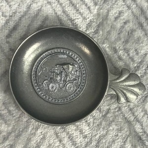 Puede incluir: Un pequeño plato de color peltre con un asa decorativa. El plato presenta un diseño circular en relieve en el centro con una imagen de un carruaje tirado por caballos. El asa tiene un diseño estilizado en forma de hoja.