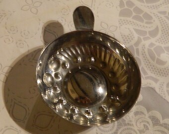 Peut inclure: Un petit moule en métal argenté avec une poignée. Le moule a une forme de bol rond avec un motif décoratif de cercles et de lignes en relief. La poignée est une petite languette plate. Le moule est posé sur une surface blanche à motifs.