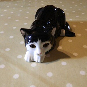 以下が含まれることがあります： 黒と白の猫の陶器製の置物。猫は光沢のある仕上がりで、背中と頭は黒い毛、顔と足は白い毛です。猫の目は茶色で、鼻はピンク色です。