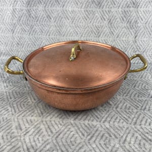 Puede incluir: Una olla de cocina de color cobre con tapa y dos asas de latón. La olla tiene forma redonda y una tapa con asa de latón. Las asas son curvas y están unidas a los lados de la olla.