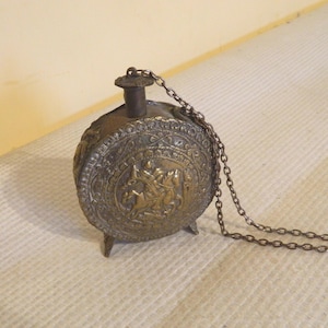 Könnte beinhalten: Eine runde, antik wirkende Metallflasche mit einem geprägten Relief einer Figur zu Pferd innerhalb eines dekorativen Randes. Die Flasche hat einen kleinen Hals, eine kurze Kette am Verschluss und steht auf zwei kleinen Füßen mit dunkler Patina.