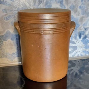 May include: A cylindrical brown stoneware crock with a matching lid and two side handles. The exterior features a smooth, matte finish with subtle horizontal ridges near the top rim, providing a simple and functional design for storage purposes.