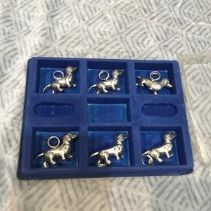 May include: A set of six silver-colored dachshund charms, each with a small loop at the top. The charms are arranged in a blue velvet-lined tray with individual compartments. The dachshunds are in a standing pose.