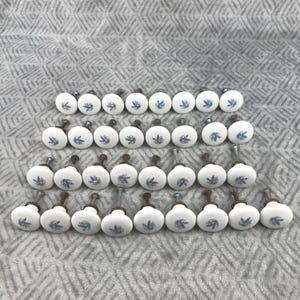 May include: A collection of white ceramic cabinet knobs with a floral design in blue and green. The knobs have a round shape and are arranged in rows on a textured gray surface. Each knob has a metal screw for installation.