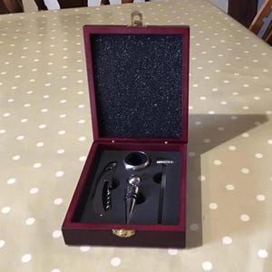 May include: A dark wood wine accessory set in a hinged box with a black foam interior. The set includes a silver-toned corkscrew, a circular bottle ring, a metal bottle stopper, and a small silver-toned tool, all arranged in custom cutouts.
