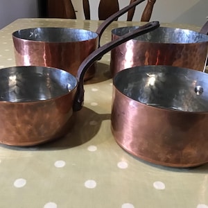 May include: Four copper saucepans with hammered exteriors and silver interiors are arranged on a light green tablecloth with white polka dots. Each pan has a long, dark handle. The pans vary slightly in size.