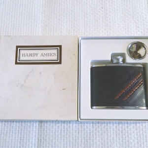 Pode incluir: Uma petaca de metal da marca Hardy Amies com revestimento de couro escuro com duas faixas diagonais costuradas em castanho, apresentada numa caixa branca com um funil de metal. A tampa da caixa exibe o texto Hardy Amies dentro de uma moldura.
