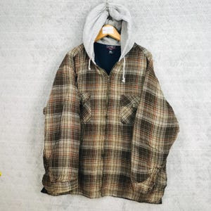 Puede incluir: Chaqueta con capucha a cuadros marrón, beige y blanco con capucha gris y cremallera completa. La chaqueta tiene dos bolsillos delanteros y un interior azul oscuro. La chaqueta está colgada de una percha de madera.