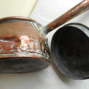 May include: A vintage copper saucepan with a long handle and a matching circular lid. The exterior of the pan shows a weathered, textured surface with visible oxidation and patina, while the interior of the lid is dark and metallic.