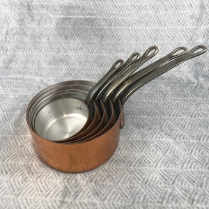 May include: A set of copper saucepans with silver handles. The saucepans are nested inside each other, showcasing their varying sizes. The copper exterior contrasts with the silver handles and interior, creating a classic kitchenware aesthetic.