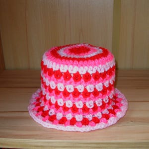 Pode incluir: Um chapéu de balde de crochê feito à mão em tons de vermelho, rosa e branco. O chapéu apresenta uma aba larga e uma coroa cilíndrica. O padrão de cores inclui círculos concêntricos no topo e fileiras alternadas de vermelho e branco.