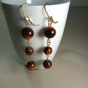 Può includere: Orecchini pendenti con tre perle marroni occhio di tigre sospese a ganci color oro. Gli orecchini hanno un tono caldo e terroso, con le perle che mostrano motivi e variazioni naturali.