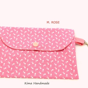 May include: A pink fabric pouch with a white floral pattern and a snap closure. The pouch has a pink strap with a silver hook. The text "M. ROSE" is printed on the top of the pouch. The text "Kima Handmade" is printed on the bottom of the pouch.