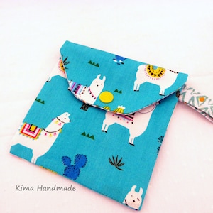 May include: A small turquoise blue fabric pouch with a white llama print. The pouch has a flap closure and a wrist strap with a key ring. The text "Kima Handmade" is printed on the bottom of the pouch.