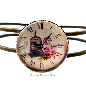 May include: A double-banded bronze bangle bracelet with a round glass cabochon featuring a vintage clock face with Roman numerals, a birdcage, and pink roses. The text "La tienda Vintage de Kima" is printed below the cabochon.