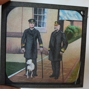 May include: Two men in navy blue uniforms and hats stand outside a building with a white dog. The men are holding canes and the dog is looking at the camera.