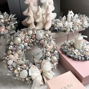 Könnte beinhalten: Ein festlicher Weihnachtskranz mit einer Hirschfigur, einer weißen Schleife und einer Auswahl an goldenen, silbernen und rosa Ornamenten. Weitere Dekorationen sind ein Kerzenkranz, dekorative Bäume und Geschenkboxen.