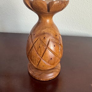 May include: A carved wooden pineapple-shaped candle holder. The candle holder is brown with a diamond pattern carved into the body and a flower-like top. The base is round and the overall design is tropical.