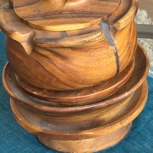May include: A set of four wooden bowls with a lid. The bowls are stacked on top of each other and are made of light brown wood. The lid has a unique design with a raised centre.
