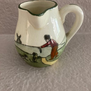 May include: A small, white ceramic creamer with a green rim and handle. It features a hand-painted scene of a person walking with two dogs in a green field.