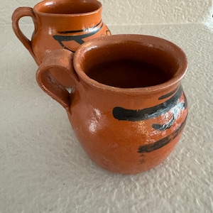 May include: Two terracotta mugs with black painted designs. The mugs have a rounded body and a handle.
