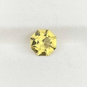 May include: A faceted, octagonal gemstone with a pale yellow hue. The gem has multiple facets that reflect light, creating a sparkling effect. The gemstone is set against a white background.