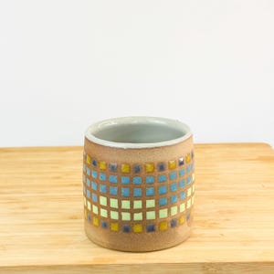May include: A ceramic mug with a mosaic tile design. The mug is cylindrical with a tan base and a pattern of blue, yellow, and green squares. The interior is a light grey colour.