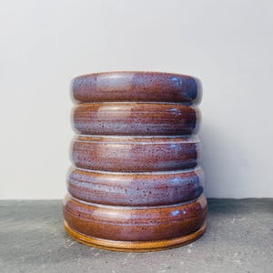 May include: A handmade ceramic vase with a stacked, cylindrical design. The vase has a rich, earthy colour palette of brown, purple, and blue hues. The exterior has a glossy finish, and the base is a lighter shade of brown.