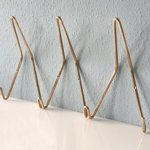 Può includere: Tre ganci da parete in metallo color oro dal design minimalista. Ogni gancio ha una parte superiore curva e un gancio rivolto verso il basso. I ganci sono montati su una parete strutturata azzurra.
