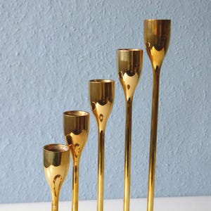Puede incluir: Cinco candelabros de metal dorado de diferentes alturas. Los candelabros son todos de forma cil&iacute;ndrica con una parte superior acampanada.