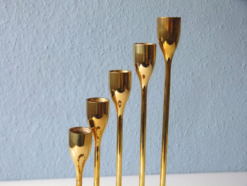 Puede incluir: Cinco candelabros de metal dorado de diferentes alturas. Los candelabros son todos de forma cil&iacute;ndrica con una parte superior acampanada.