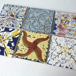 Op de afbeelding: Zes vierkante keramische tegels met handgeschilderde ontwerpen. De tegels tonen verschillende patronen, waaronder een zeester, abstracte figuren en bloemmotieven. Het kleurenpalet omvat blauw, geel, rood en groen, waardoor een levendige en artistieke weergave ontstaat.