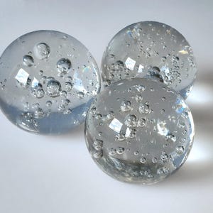 Könnte beinhalten: Drei transparente Glaskugeln mit inneren Blasen. Die Kugeln sind klar, so dass Licht durchscheinen kann, und sind auf einer weißen Oberfläche angeordnet. Die Blasen variieren in der Größe und verleihen den Glaskugeln visuelles Interesse.