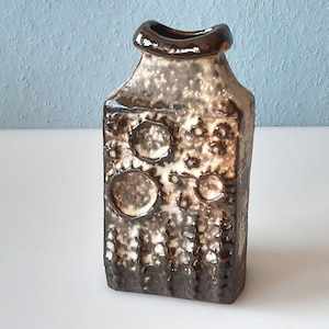 Pode incluir: Um vaso retangular de cerâmica com uma superfície texturizada e uma borda marrom escura. O vaso tem um padrão salpicado e manchado em tons de marrom e bege, com desenhos circulares e ovais em relevo. O vaso tem cerca de 20 cm de altura.