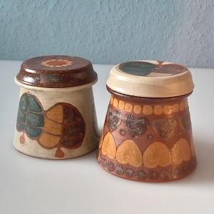 Könnte beinhalten: Zwei Keramikbehälter mit Deckeln. Ein Behälter ist cremefarben mit einem türkis-braunen Design. Der andere Behälter ist braun mit einem herzförmigen Muster. Beide Behälter haben braune Deckel.