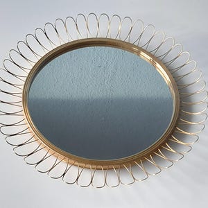 May include: A round, gold-toned mirror with a decorative wire frame. The mirror has a simple, elegant design, with a circular reflective surface and a sunburst-style frame. The frame is made of thin, curved wires that create a delicate, openwork pattern.