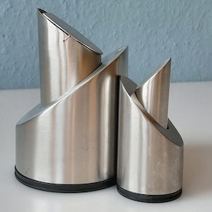 May include: Two stainless steel salt and pepper shakers with a modern, angular design. The shakers have a brushed metal finish and black rubber bases. The larger shaker has a hinged lid. The background is a light blue wall.