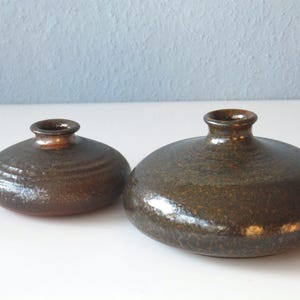 May include: Two brown ceramic vases with a speckled glaze. The smaller vase is round and has a slightly wider opening than the larger vase. The larger vase is wider and has a more rounded opening.