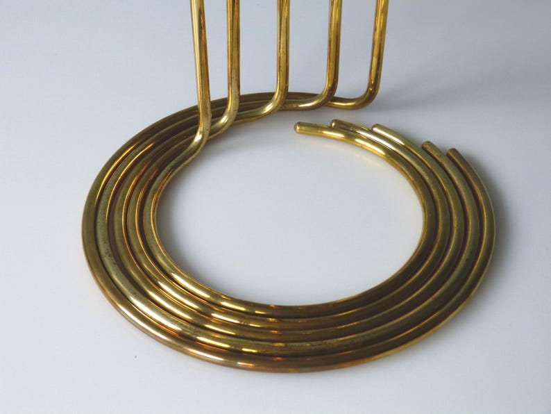 Puede incluir: Un candelero de metal dorado con una base circular y cinco brazos verticales. La base est&aacute; hecha de m&uacute;ltiples anillos conc&eacute;ntricos.