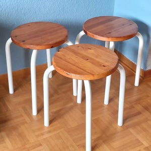 Può includere: Tre sgabelli con sedili rotondi in legno marrone medio e quattro gambe in metallo bianco ciascuno. Gli sgabelli sono disposti in gruppo, caratterizzati da una finitura in legno lucido con venature visibili sui piani circolari e telai tubolari bianchi.