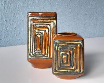 Può includere: Due vasi in ceramica marrone con un motivo geometrico quadrato nei toni del marrone, nero e crema. I vasi sono di diverse dimensioni, con una forma rettangolare e una forma arrotondata. Lo sfondo è una parete azzurra.
