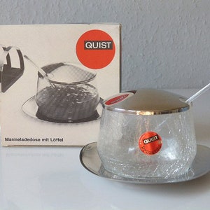May include: A vintage glass marmalade jar with a silver lid and spoon, resting on a silver saucer. The jar has a crackled texture and a red "QUIST" sticker. A box with the "QUIST" logo is in the background.