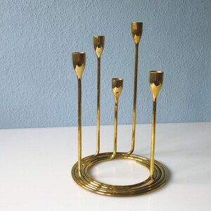 Puede incluir: Un candelero de metal dorado con cinco brazos, cada uno con una copa redonda para sostener una vela. La base del candelero es de dise&ntilde;o espiral.