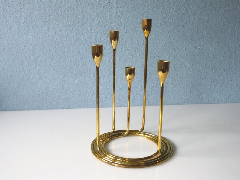 Puede incluir: Un candelero de metal dorado con cinco brazos, cada uno con una copa redonda para sostener una vela. La base del candelero es de dise&ntilde;o espiral.