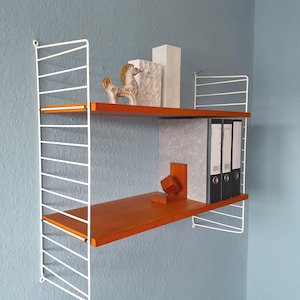 May include: A two-tiered wall-mounted shelf unit. The shelves are a warm brown with orange edging, supported by a white wire frame. The shelves display decorative items and black file folders against a blue wall.