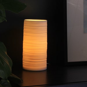 May include: A white ceramic table lamp with a textured, layered design. The lamp is lit and casting a warm glow.
